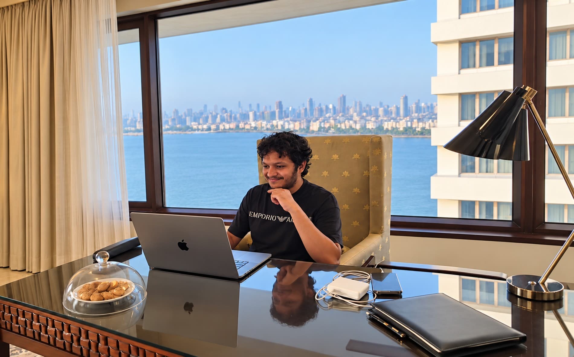 Imzzy working at a laptop by a window over the Mumbai seafront.